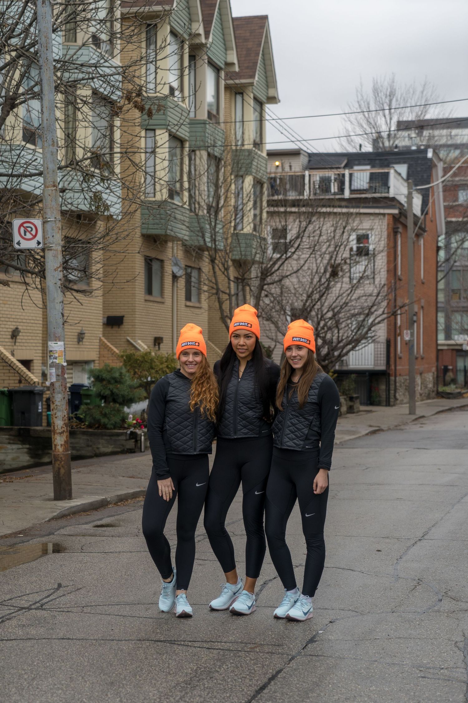 Three women in orange hats smile while standing together on a lively street.