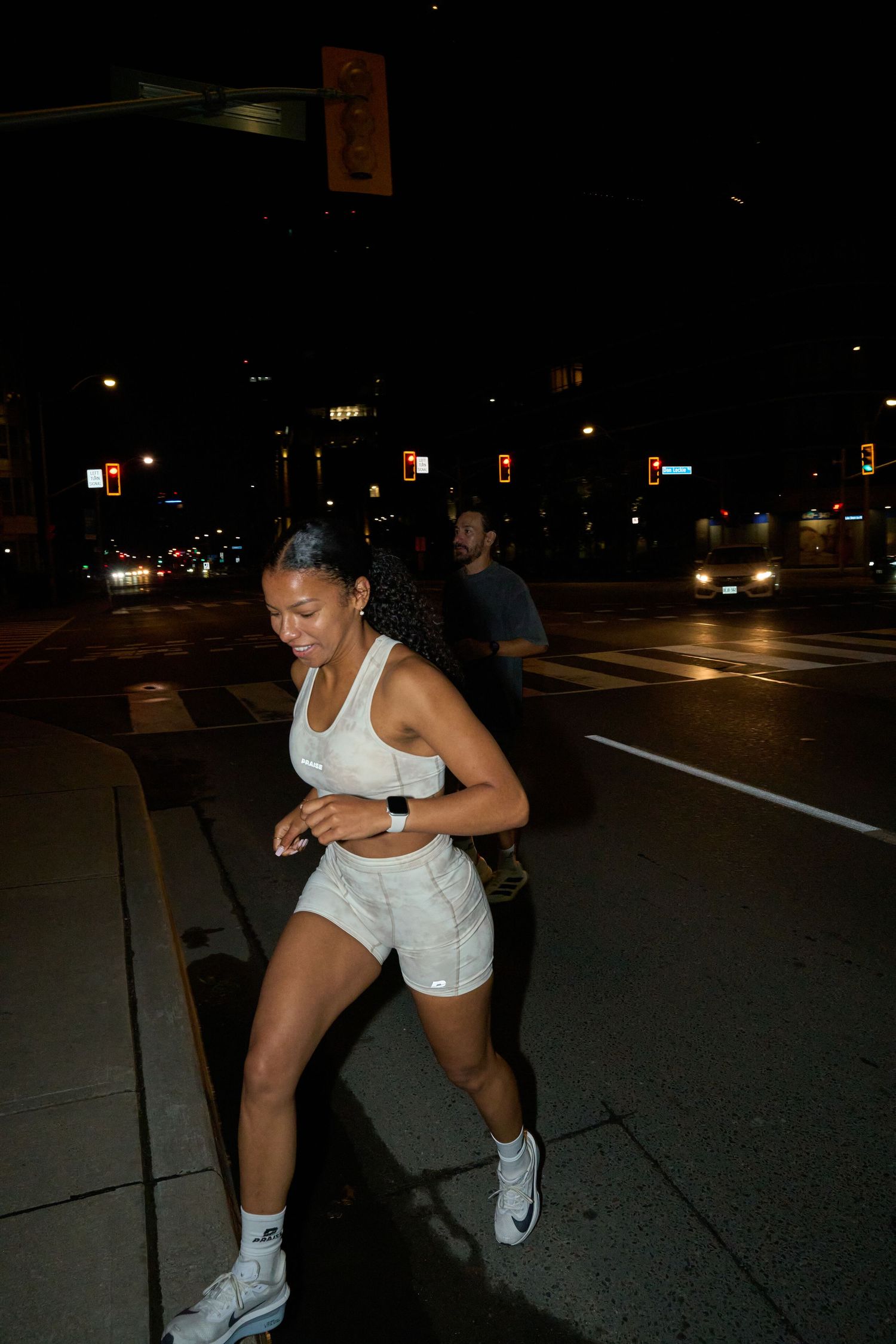 A woman in white athletic attire is situated on the roadside at night, enveloped by darkness and the subtle glow of streetlight.