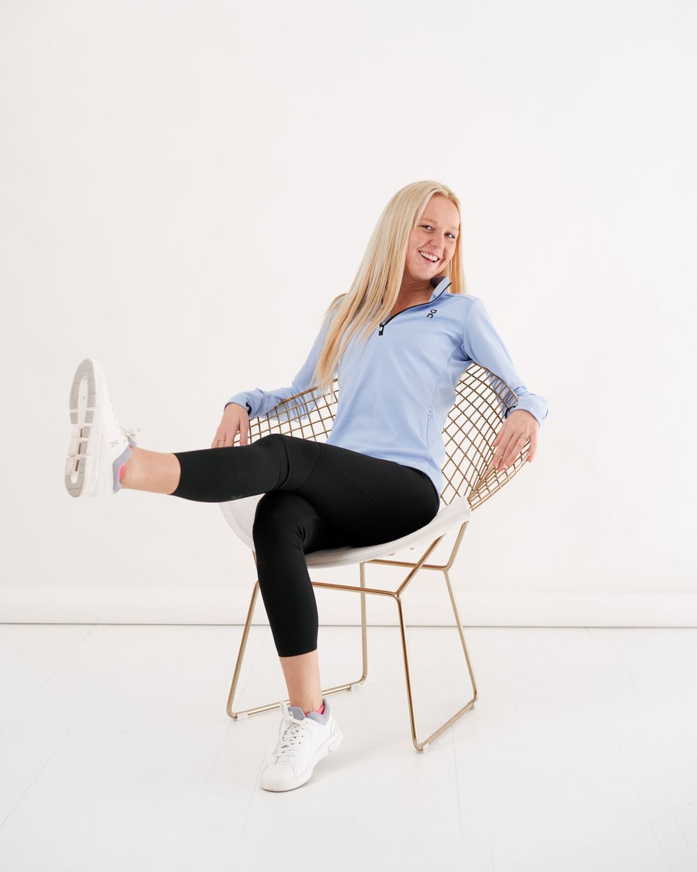 A woman in a blue shirt and black pants stands, exuding a cheerful demeanor.