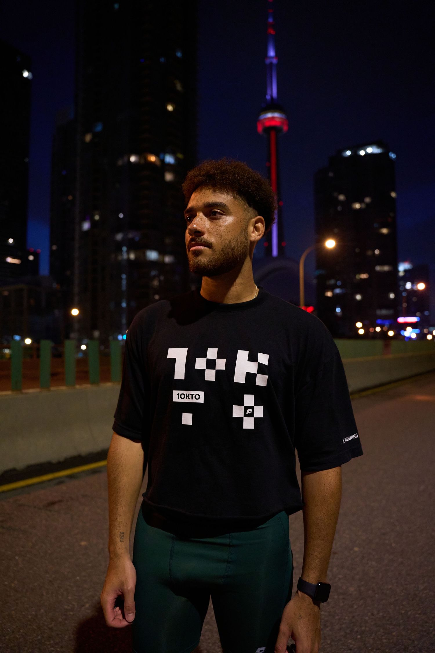 A man in black shorts and a sports shirt stands on a roadside at night, illuminated by streetlights.