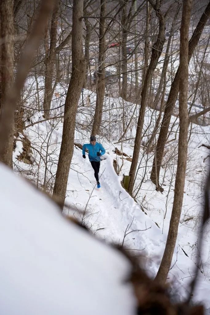 A man jogs along a snowy road next to a tranquil lake, with snow-dusted trees lining the path under a bright sky.