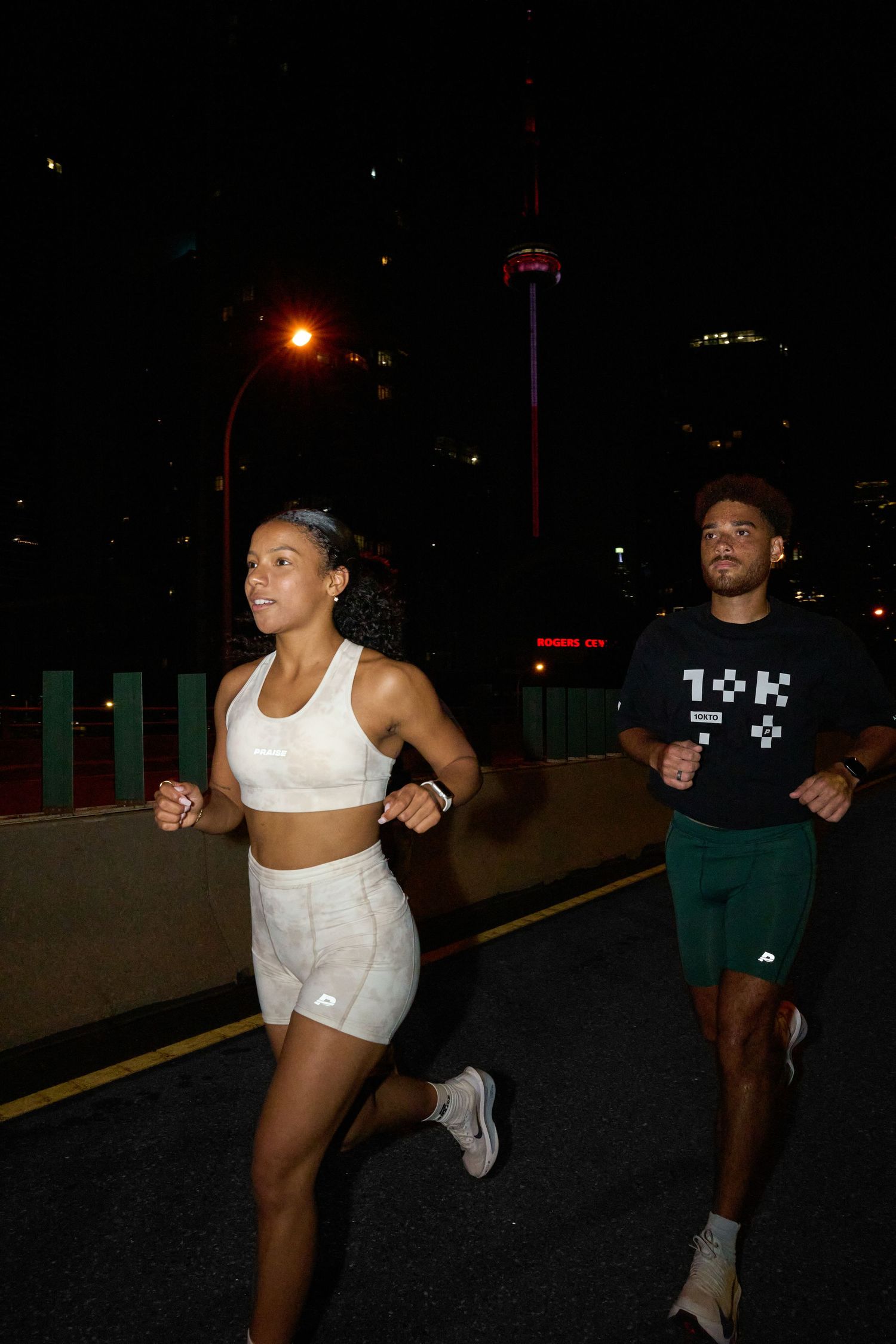 A woman dressed in white shorts and a sports bra is positioned on the roadside during the night, bathed in the light of streetlamps.