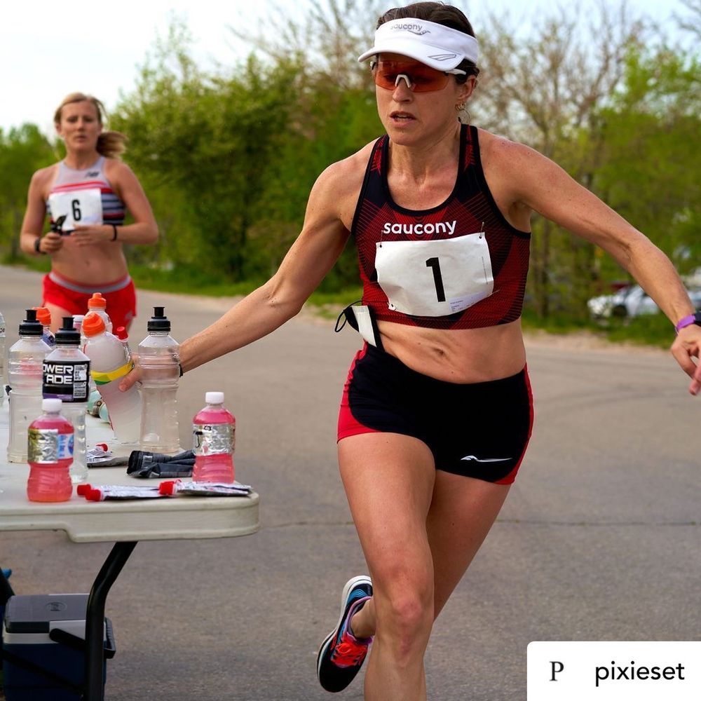 A woman sprinting in a race approaches a table filled with water bottles, showcasing her athleticism and focus.