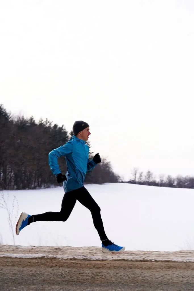 A man runs on a snowy road beside a serene lake, surrounded by snow-covered trees and a clear blue sky.