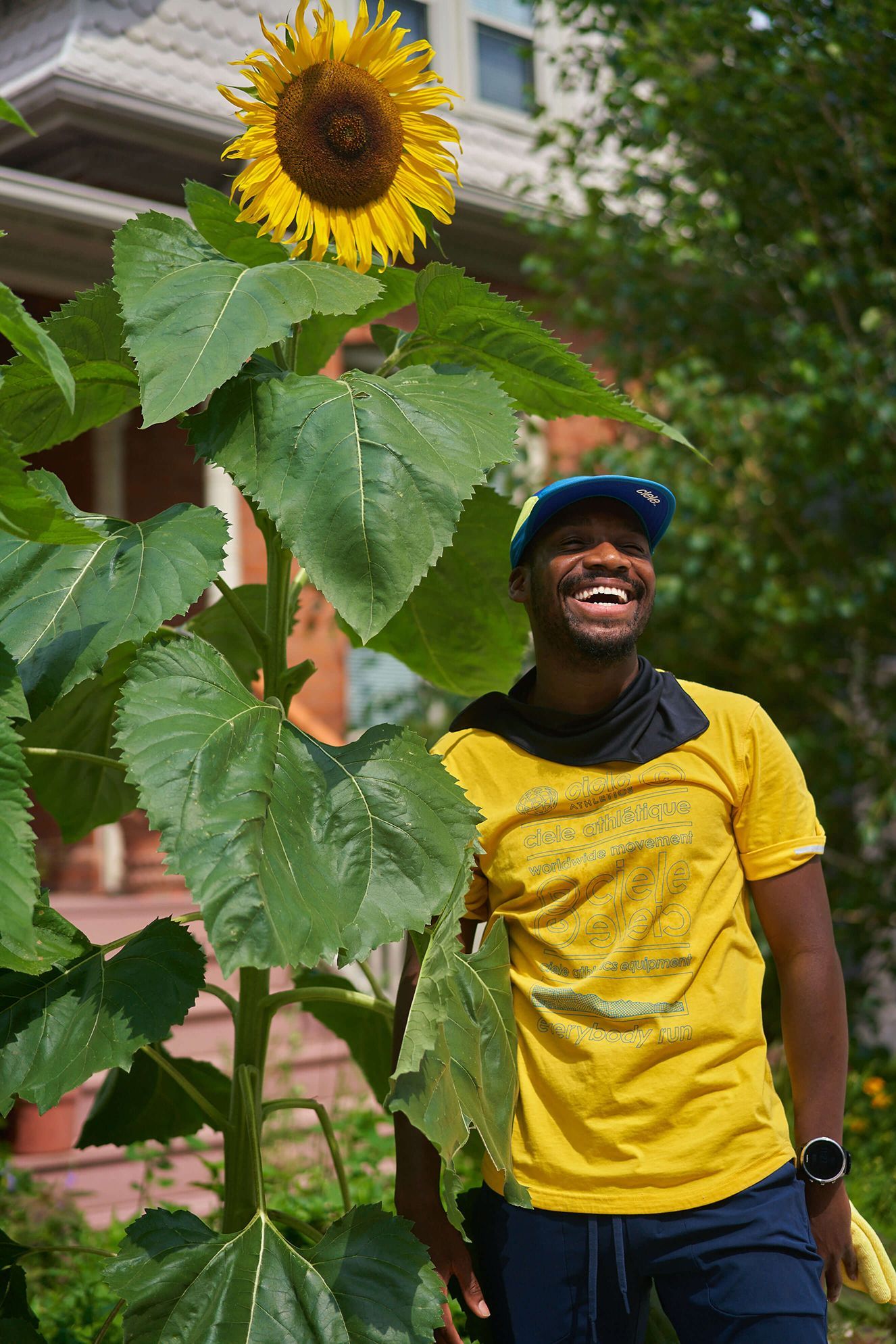 A man in a yellow shirt stands beside a large, vibrant sunflower, showcasing a cheerful outdoor scene.