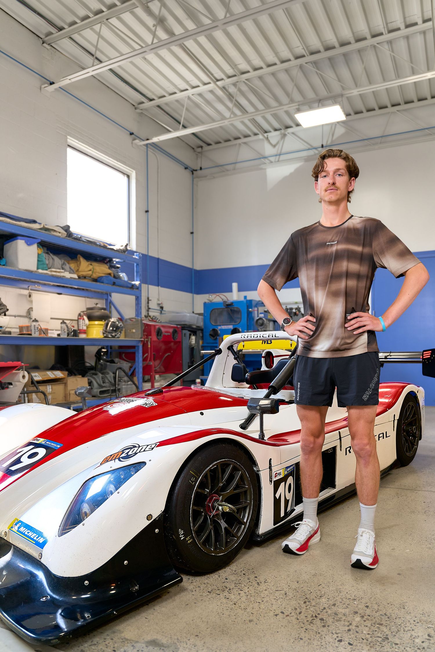 In a garage, a man poses next to a sleek race car, highlighting her passion for racing and automotive engineering.