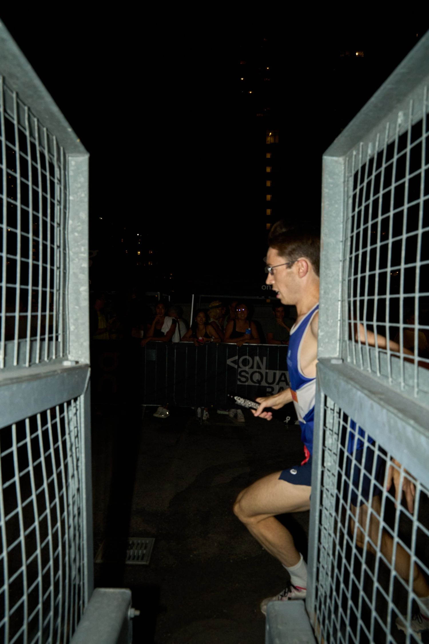 A man runner passing a metal stairway at night, with shadows cast by nearby light sources.