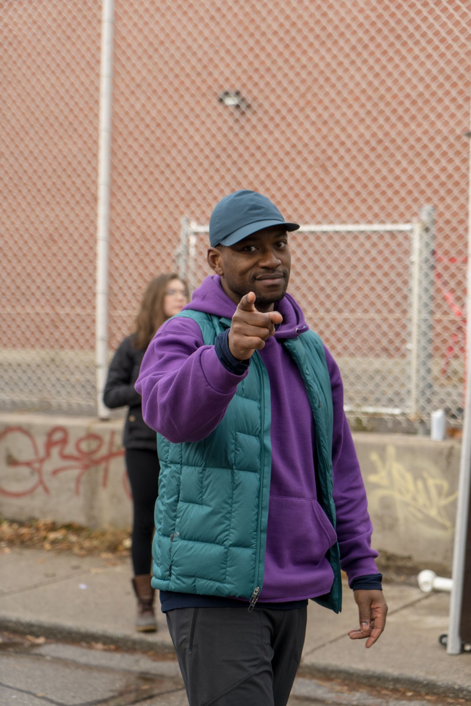 A man wearing a purple jacket and blue hat is pointing at something off-screen with a focused expression.
