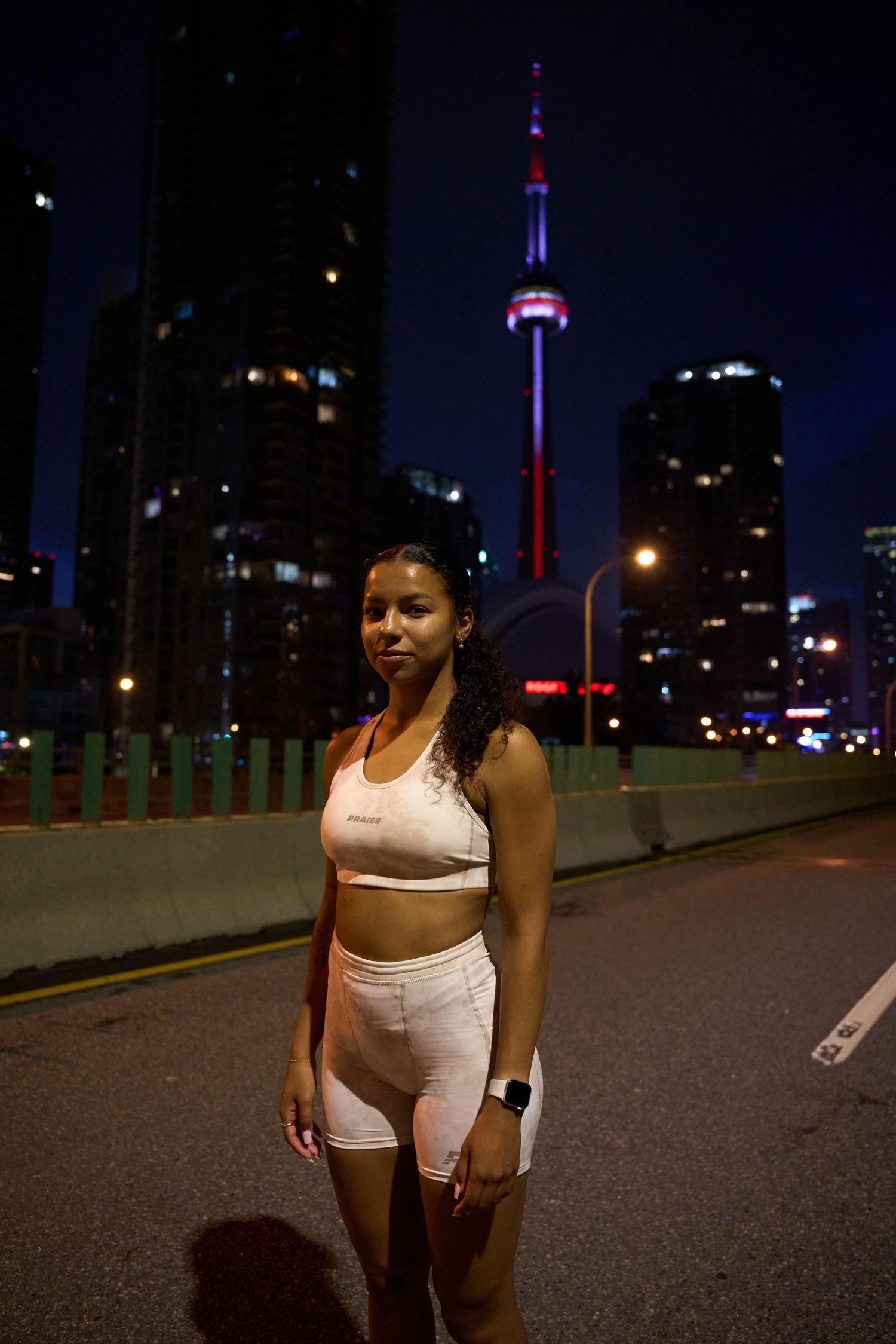 At night, a woman wearing white shorts and a sports bra stands beside a road, with streetlights casting a glow around her.