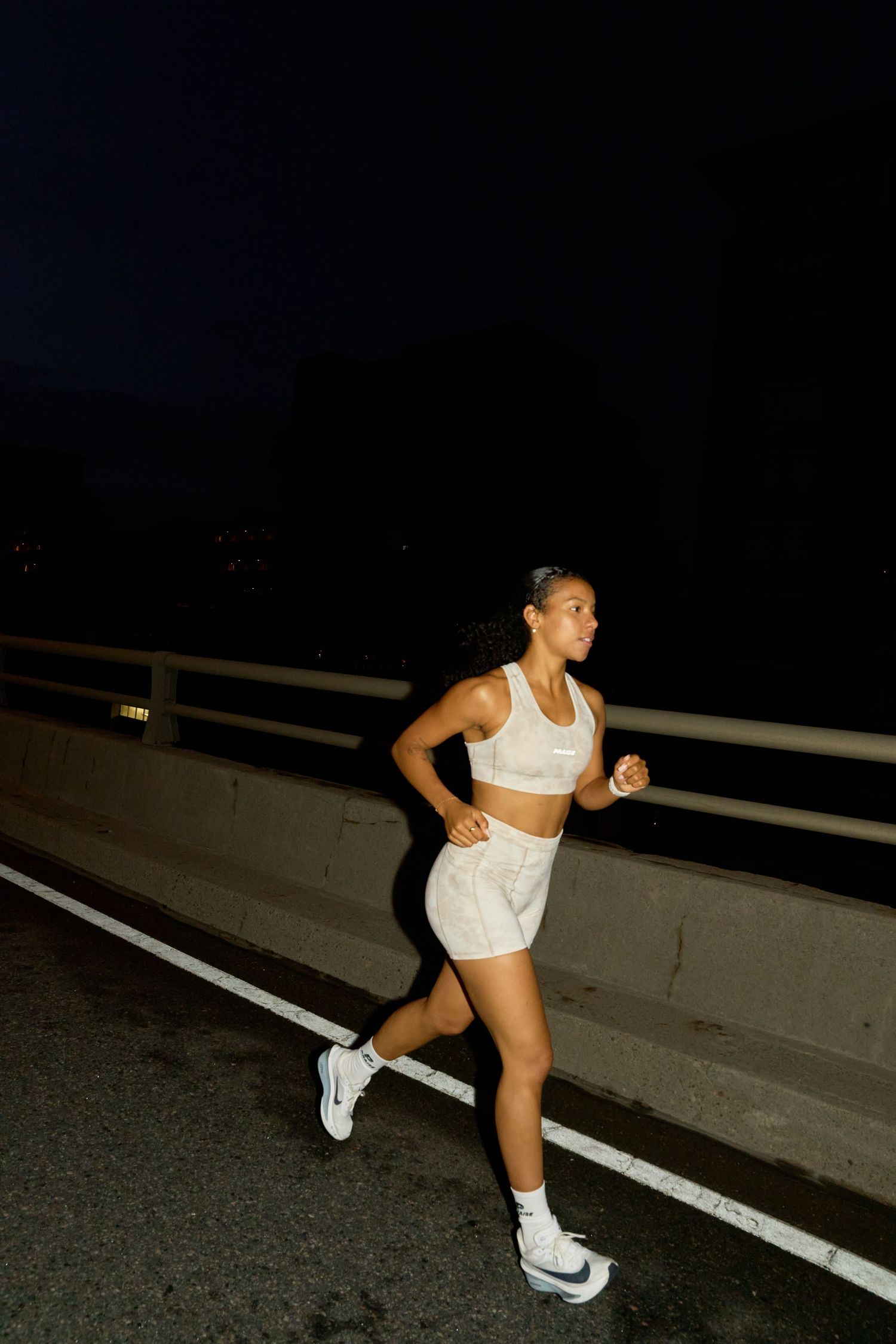 A woman in white shorts and a sports bra stands on a roadside at night, illuminated by streetlights.