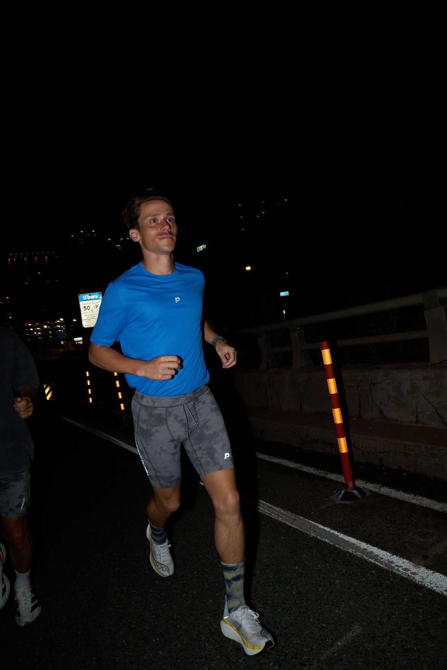 During the night, a woman clad in white shorts and a sports bra stands next to a road, with streetlights providing a soft illumination aroun