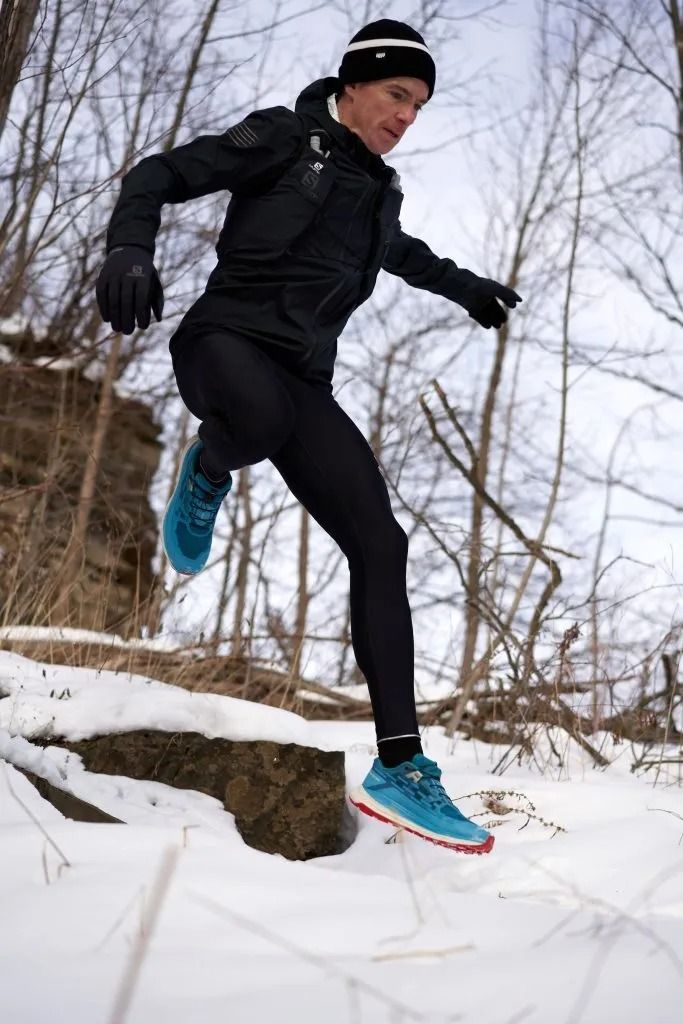 A man in winter gear runs on a snowy road adjacent to a calm lake, with snow-laden trees in the background.