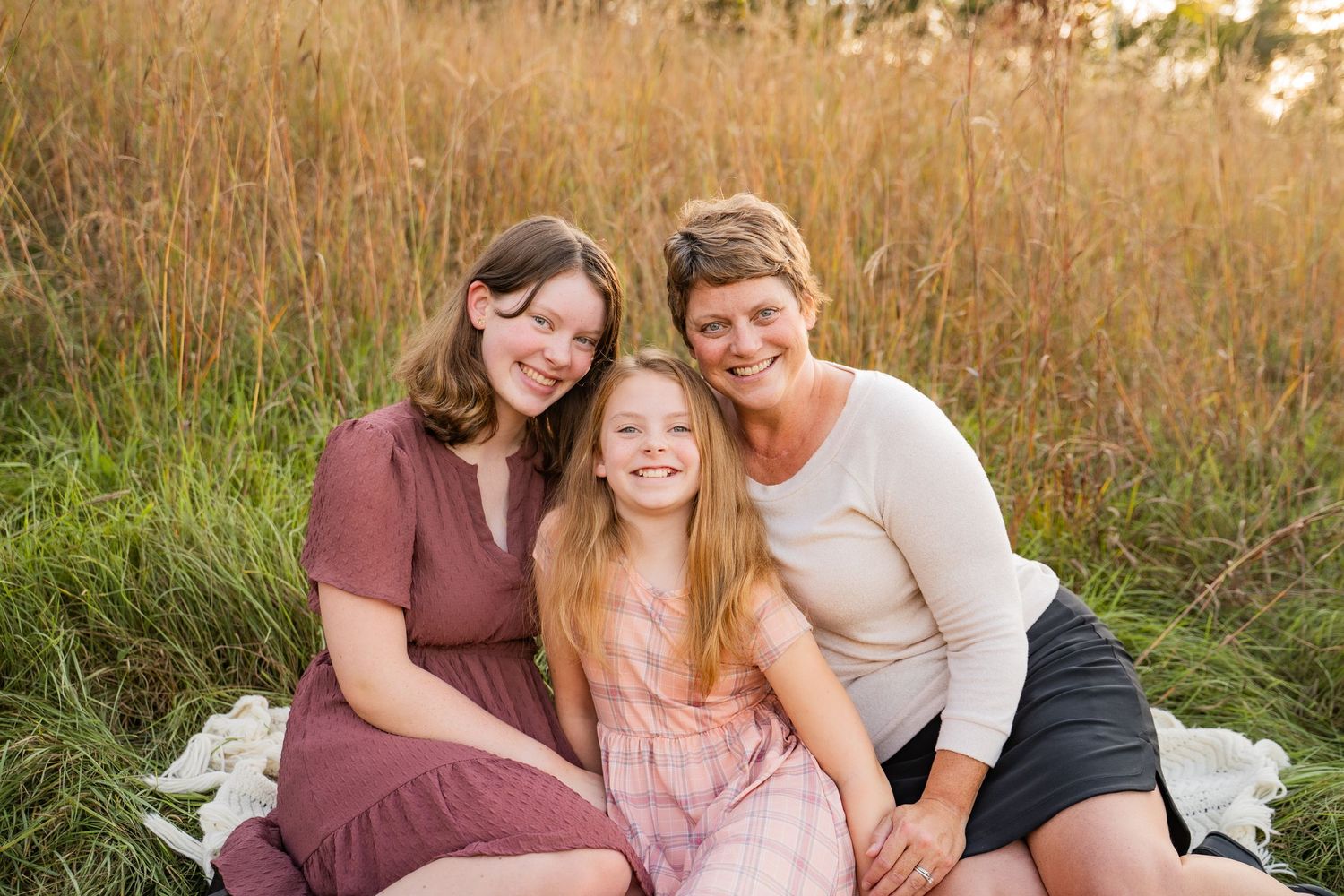 Three smiling women sitting together on a blanket in a golden grass field during a warm sunny day.