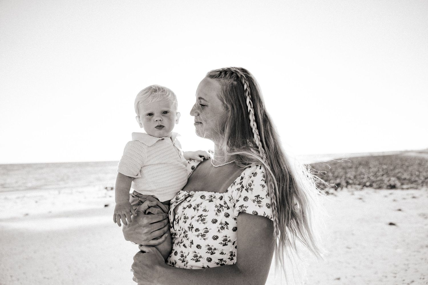Black and white photo of a mother holding her baby at the beach, both looking away from the camera.