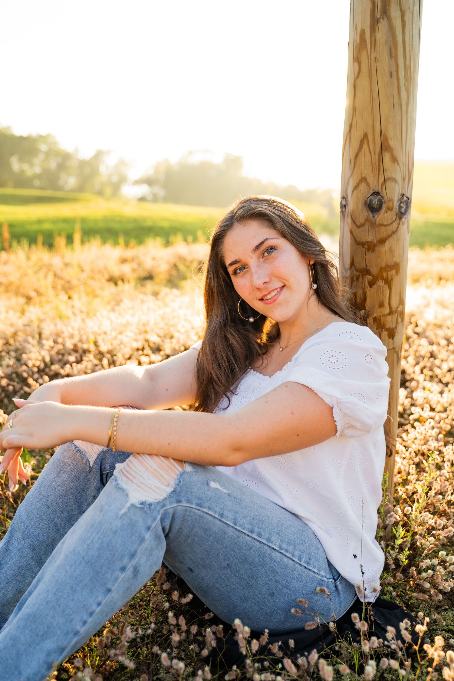 Person in white shirt and jeans for their senior session leaning against wooden post during sunset in field.