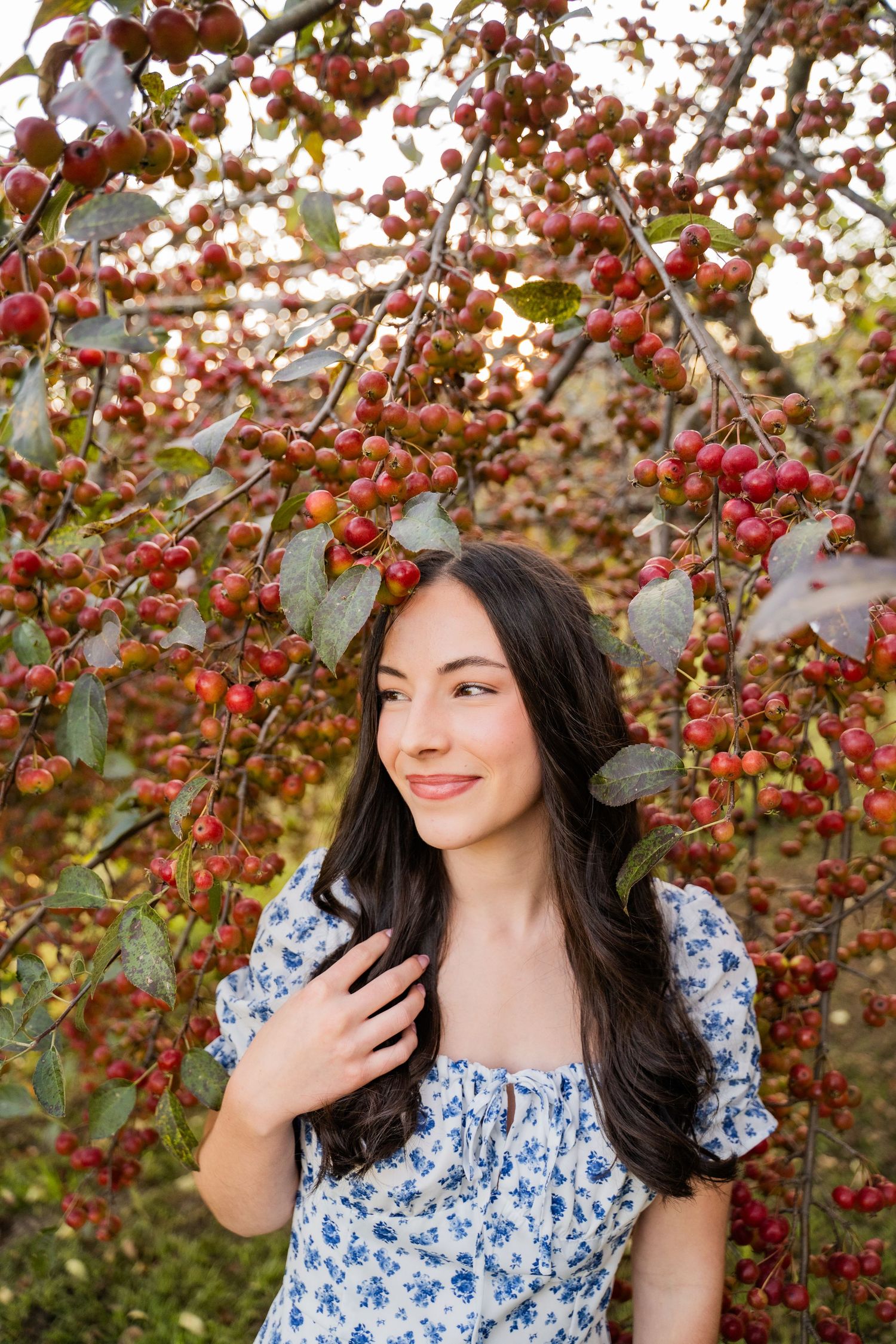 A woman in a blue floral dress poses gracefully among red berry branches.