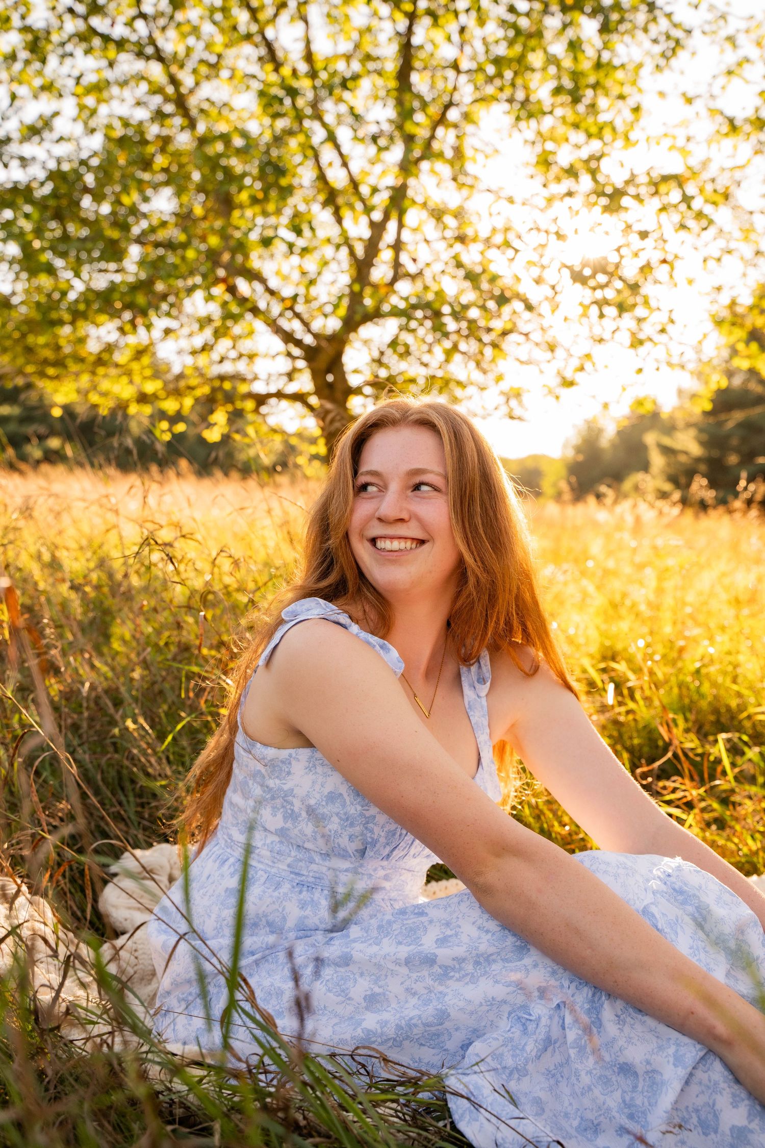 A woman in a blue floral dress relaxes in golden sunlit grass during a photo session.