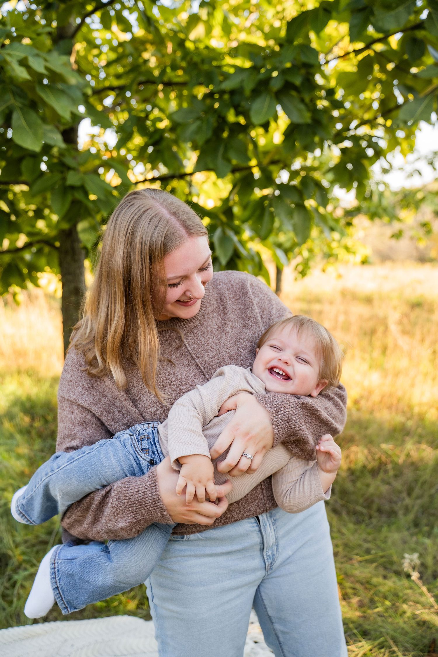 A smiling mother holds her laughing toddler outdoors in a sunny park with lush green trees in the background.
