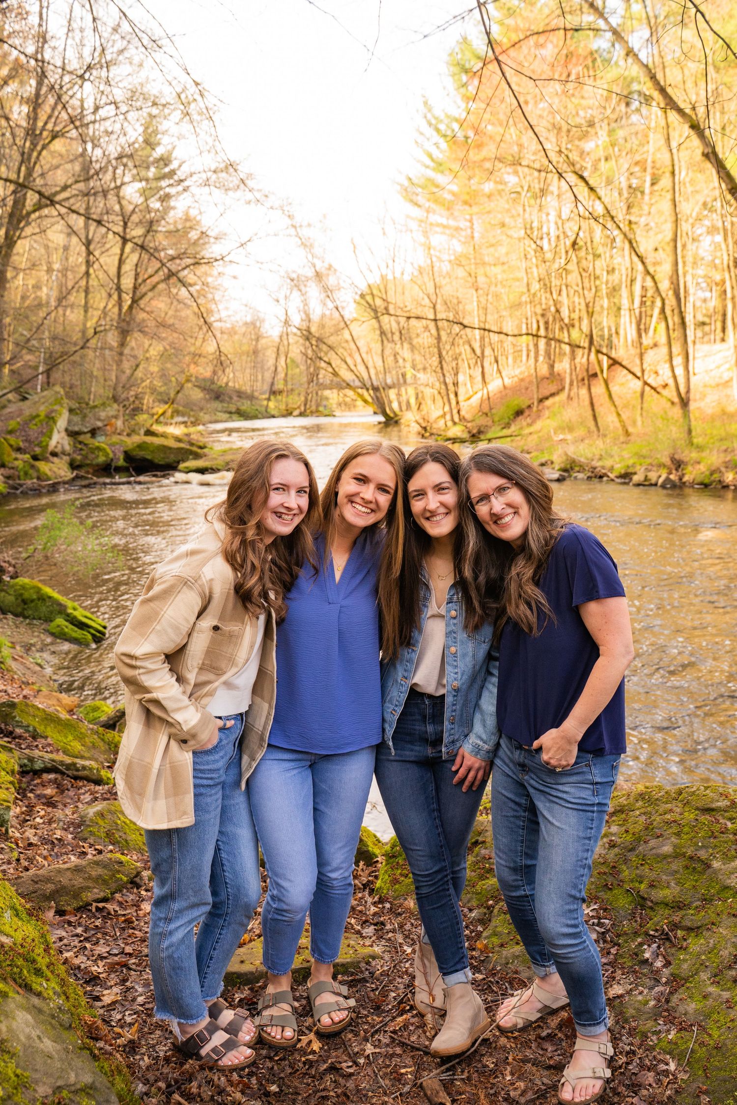 Four smiling young women pose together near a scenic creek surrounded by autumn trees in a warm outdoor setting.