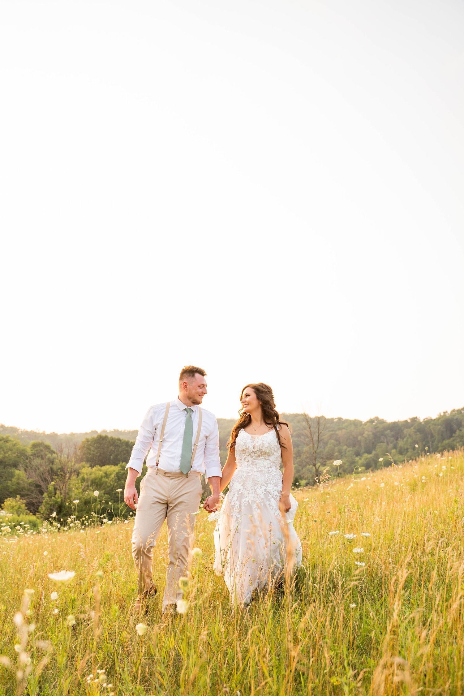 A couple walks hand in hand through a sunlit meadow of wildflowers and tall grass.
