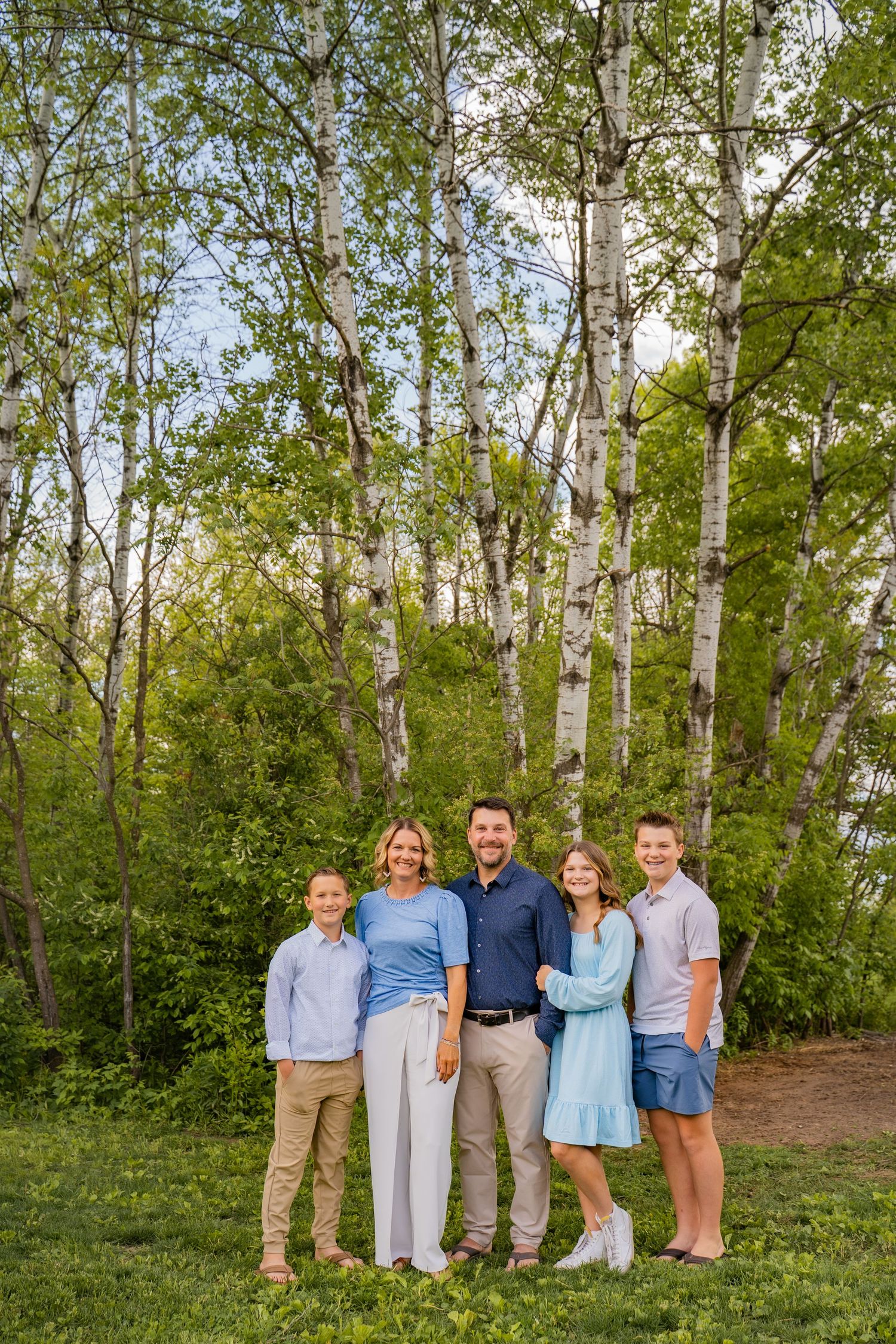 Family of five smiling outdoors among birch trees on a lush green lawn during spring.