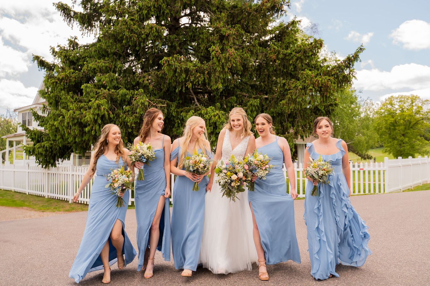A bridal party in blue dresses poses together on a sunny day with white picket fence backdrop.