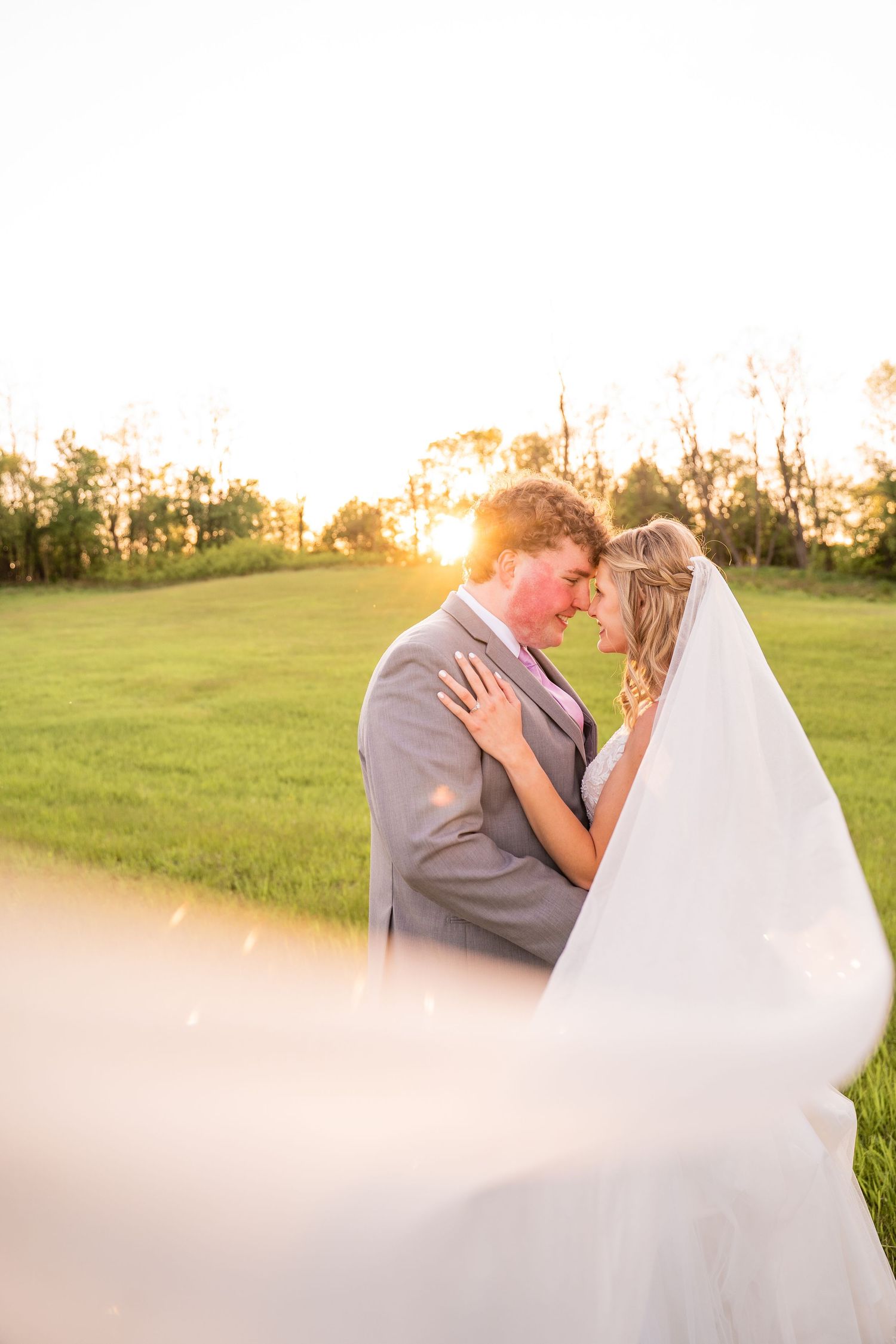 Wedding couple embracing in sunlit field with flowing veil creating dreamy effect.