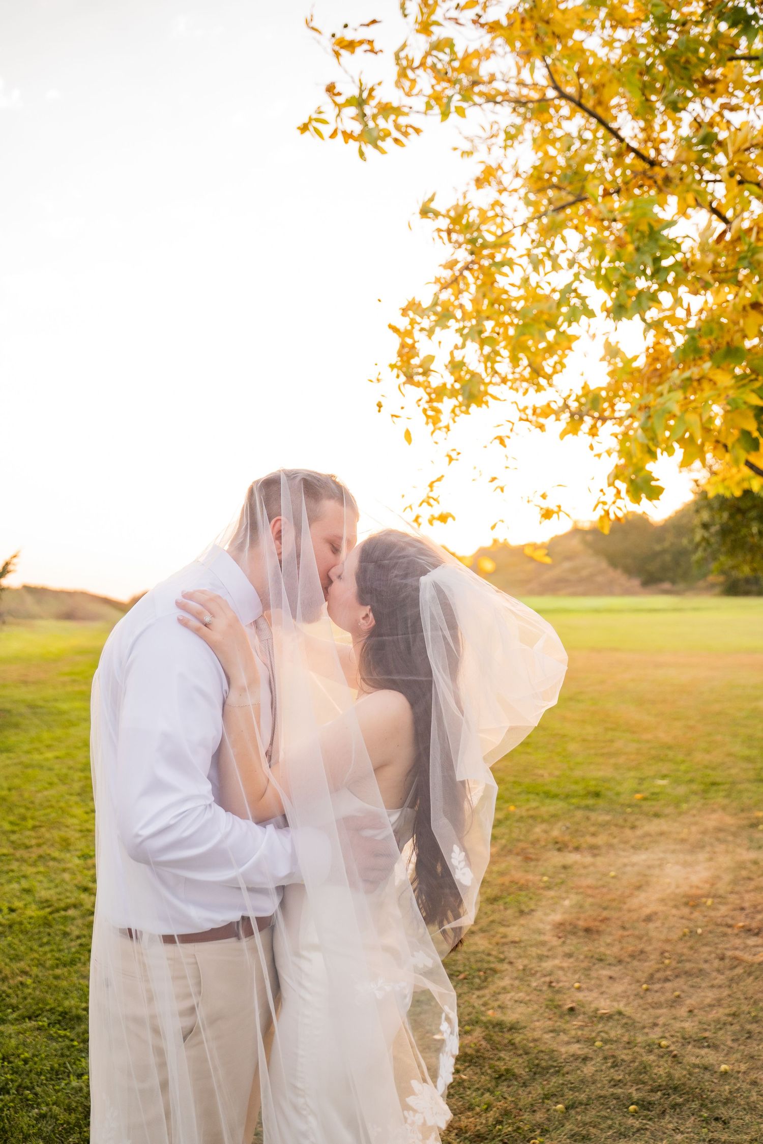 Newlyweds share romantic kiss under veil with golden autumn trees in background.