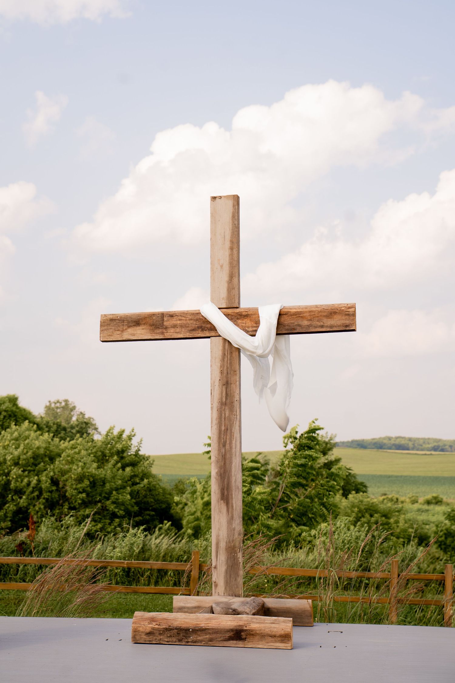 Wooden cross with white cloth draped over it against cloudy sky and green landscape.