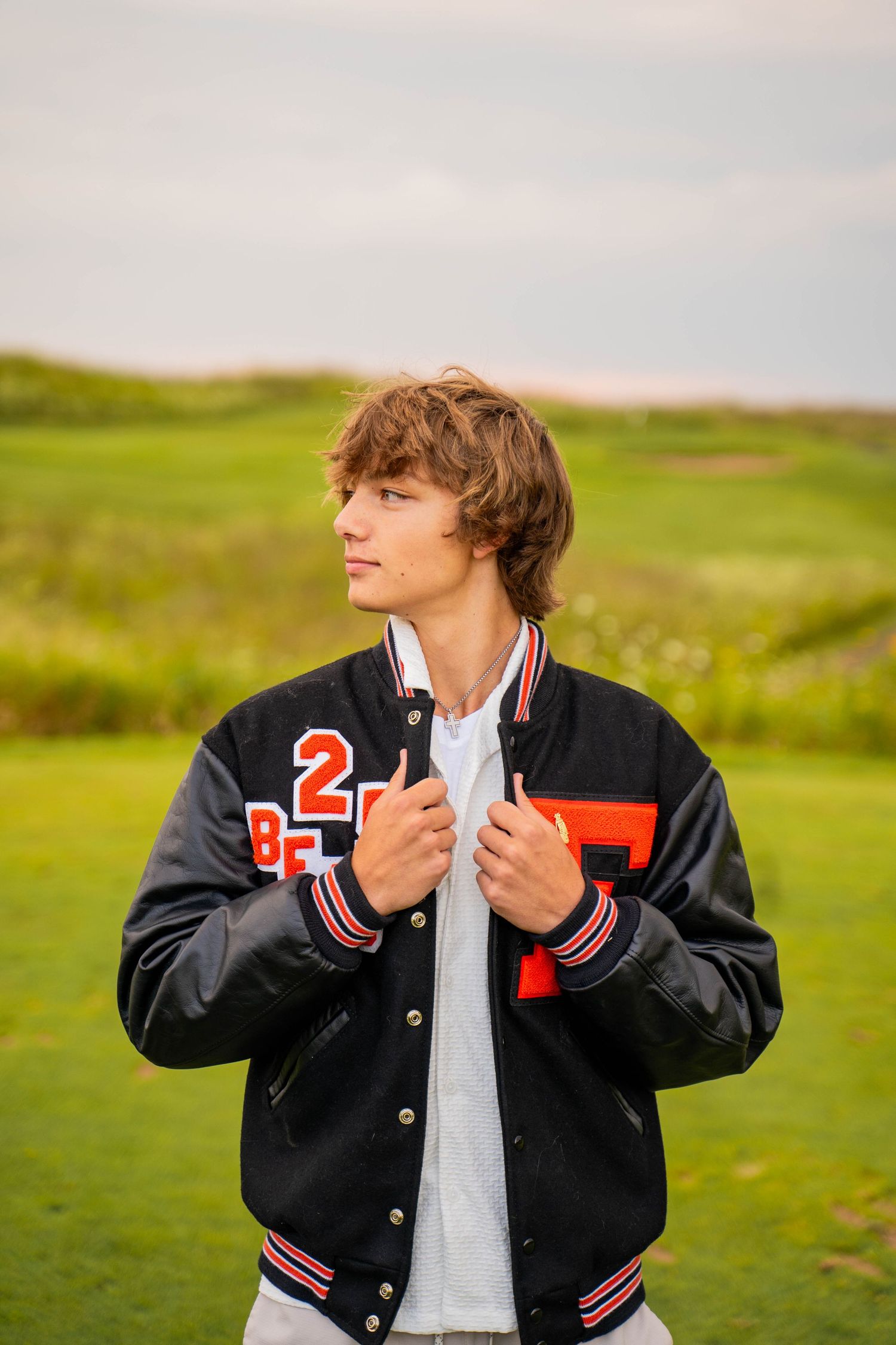 Person in varsity jacket poses against green hillside background in moody outdoor photoshoot.