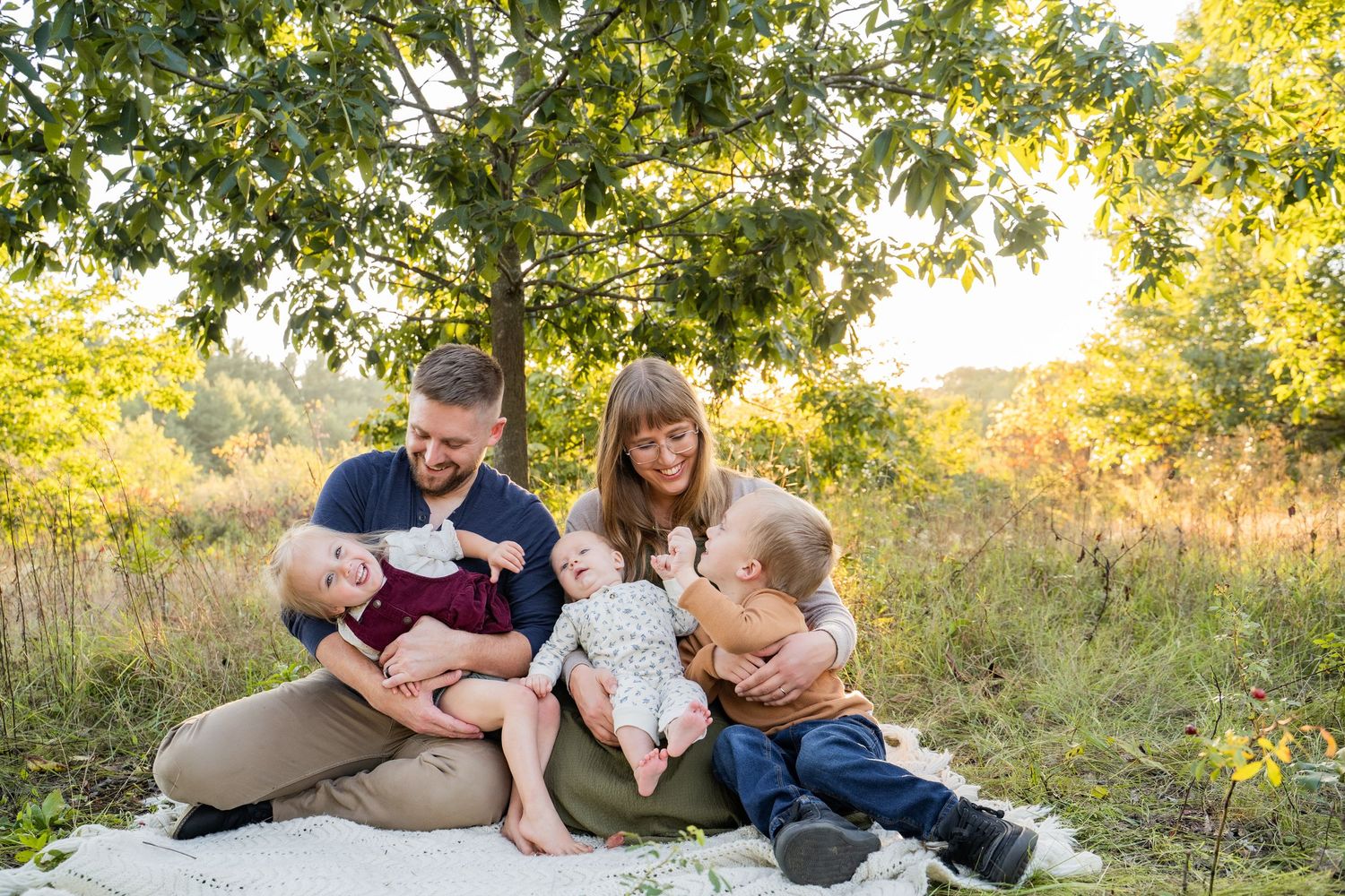 Family of five sitting on blanket under tree in golden meadow, parents holding three young children outdoors.