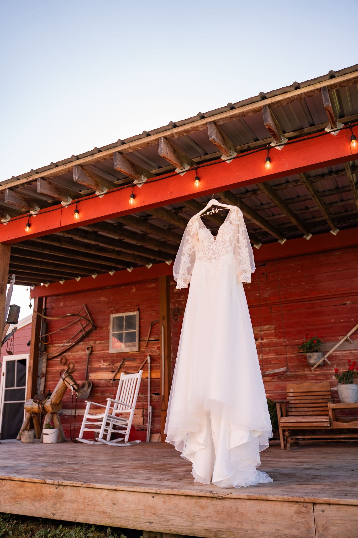 White wedding dress hanging on red barn porch with rocking chairs.