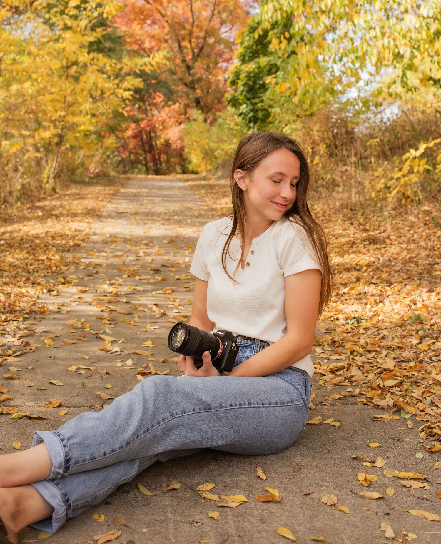 Photographer lying on autumn path with fallen leaves and camera, creating artistic angles in golden sunlight.