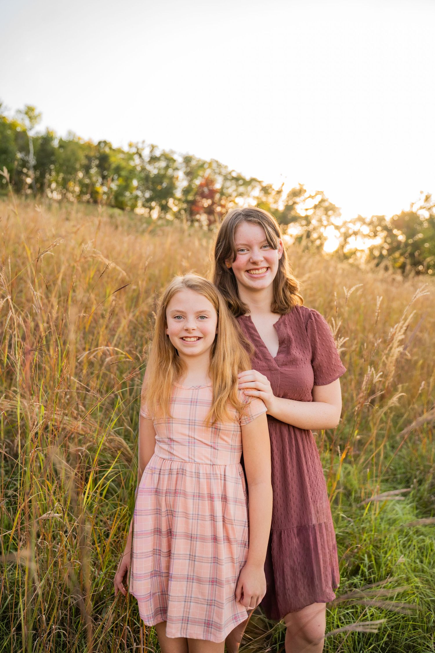 Sisters in burgundy and pink dresses stand together in a golden sunlit field of tall grass prairie at golden hour sunset in Wisconsin