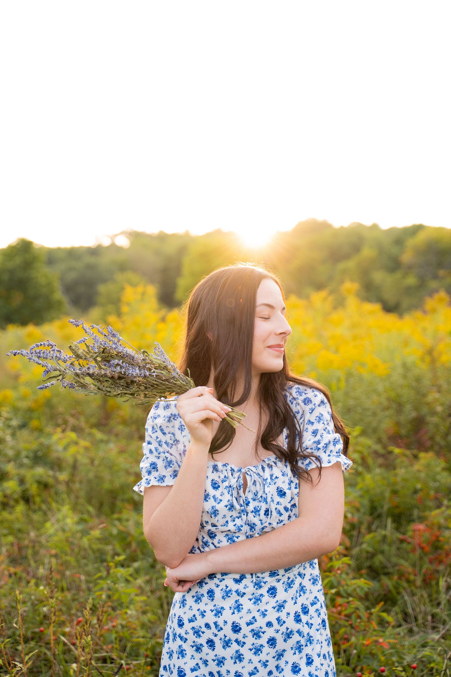 Senior portrait in blue floral dress poses joyfully in field of yellow wildflowers at sunset.