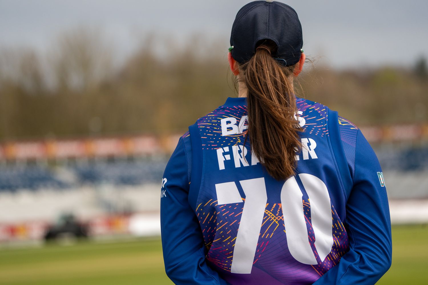 Female cricket player wearing number 70 Fraser jersey and cap, standing on cricket ground facing away from camera.