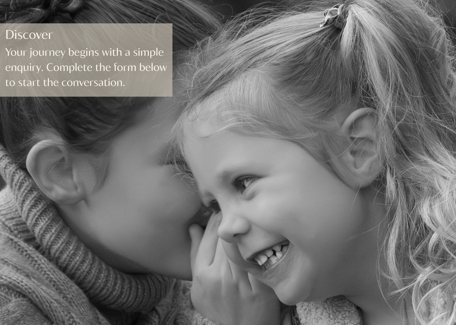 Two children share a secret and laugh together in an intimate black and white moment.