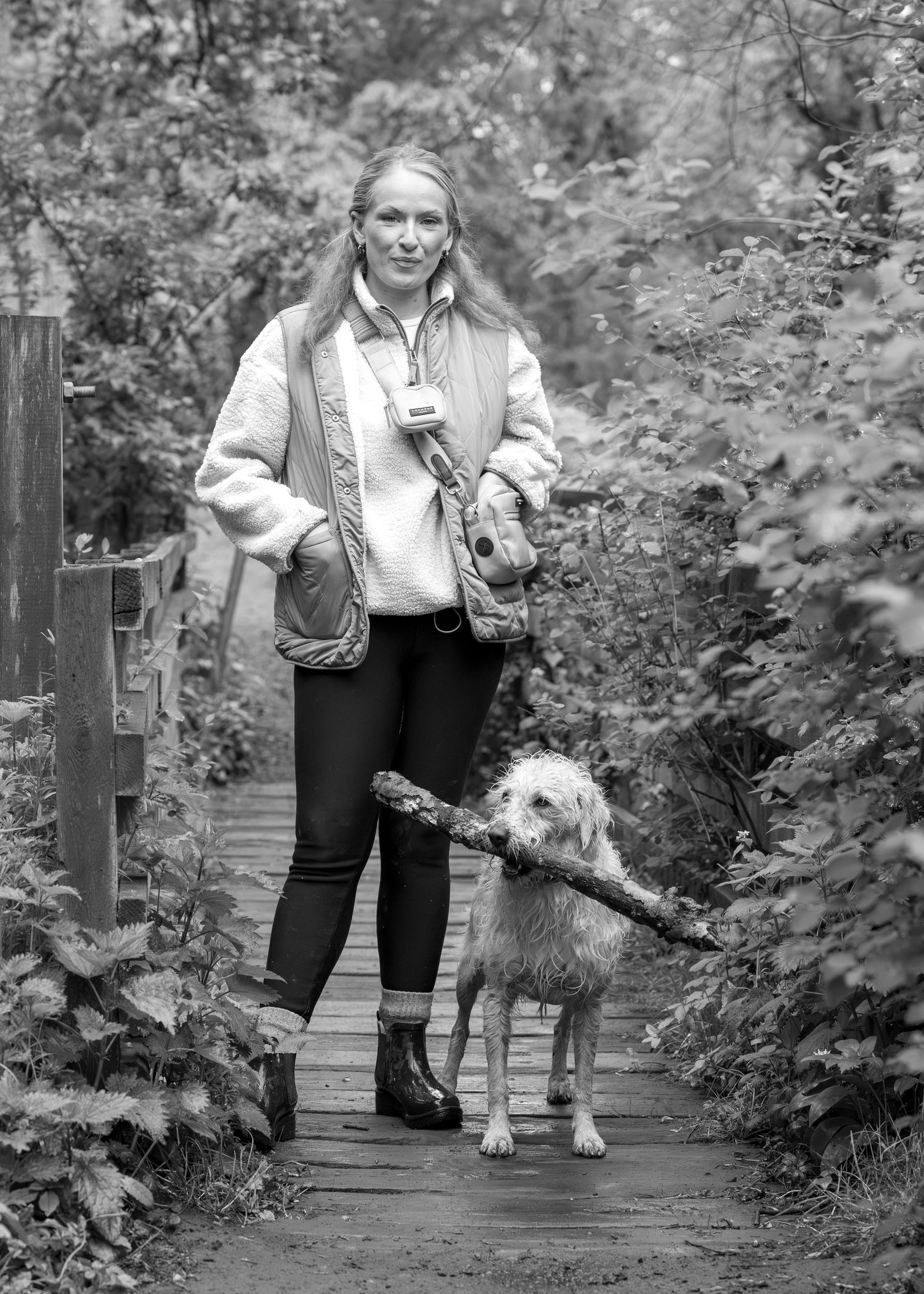 Black and white photo of a smiling woman walking a dog carrying a stick on a wooden path through lush greenery.