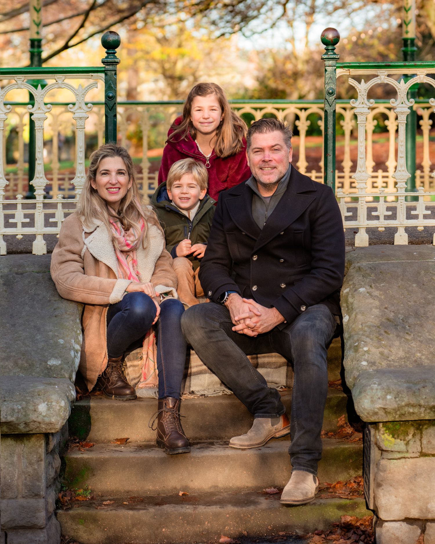 A family sits together on a stone wall during an autumn photoshoot with fallen leaves and ornate railings in the background.