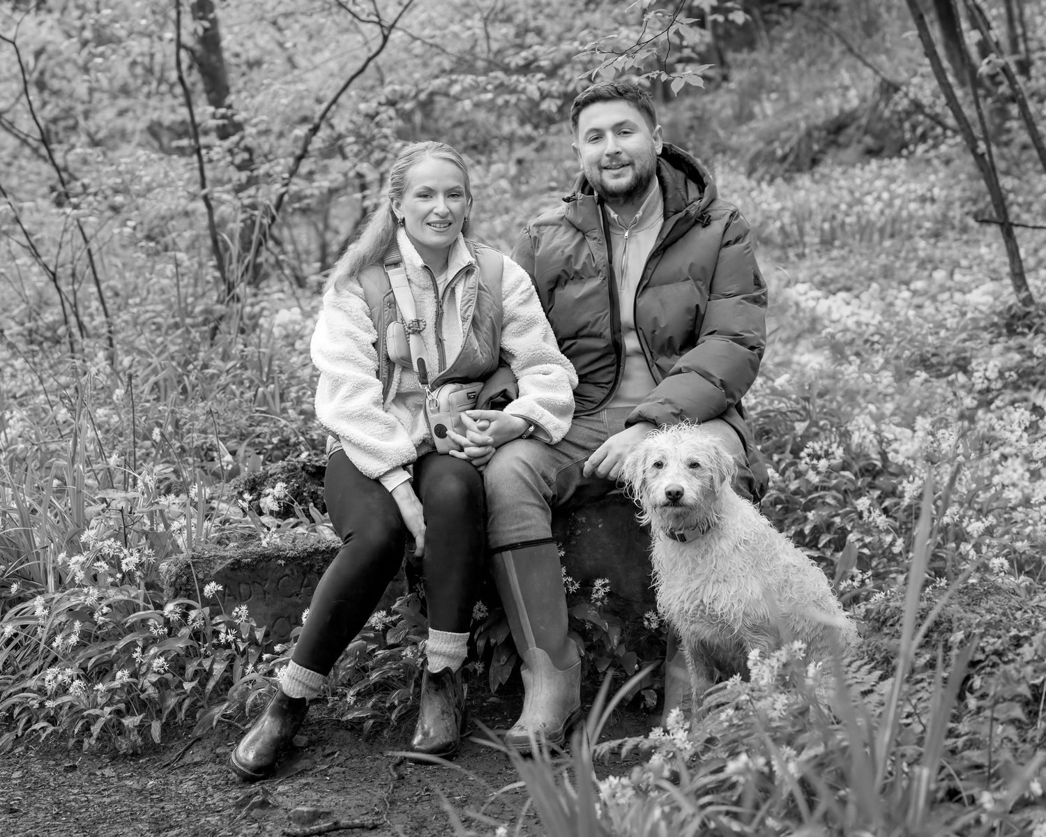 Black and white photo of a smiling couple sitting on a rock outdoors with a fluffy dog beside them.