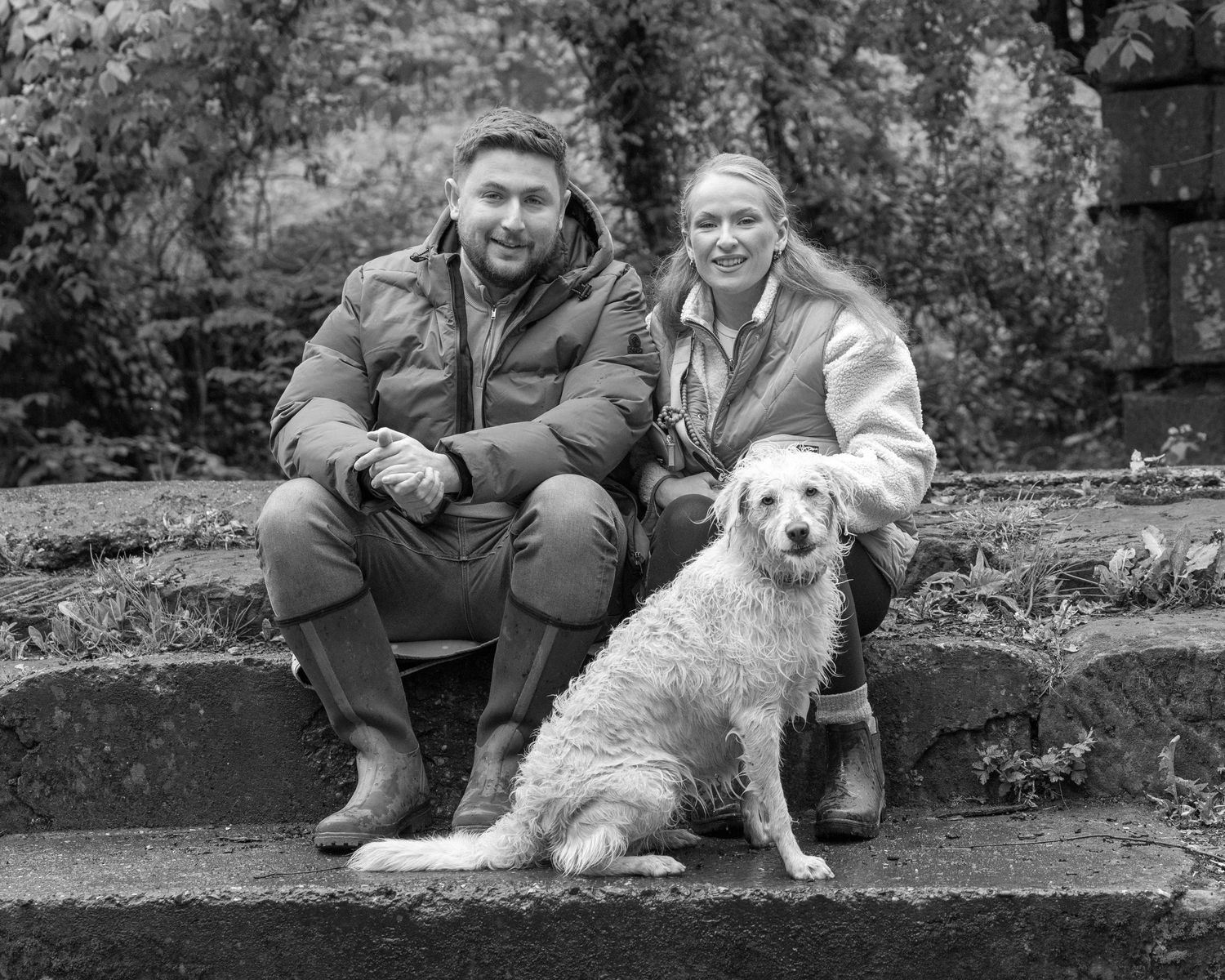 Black and white photo of a smiling couple sitting on stone steps outdoors with a fluffy golden retriever dog.