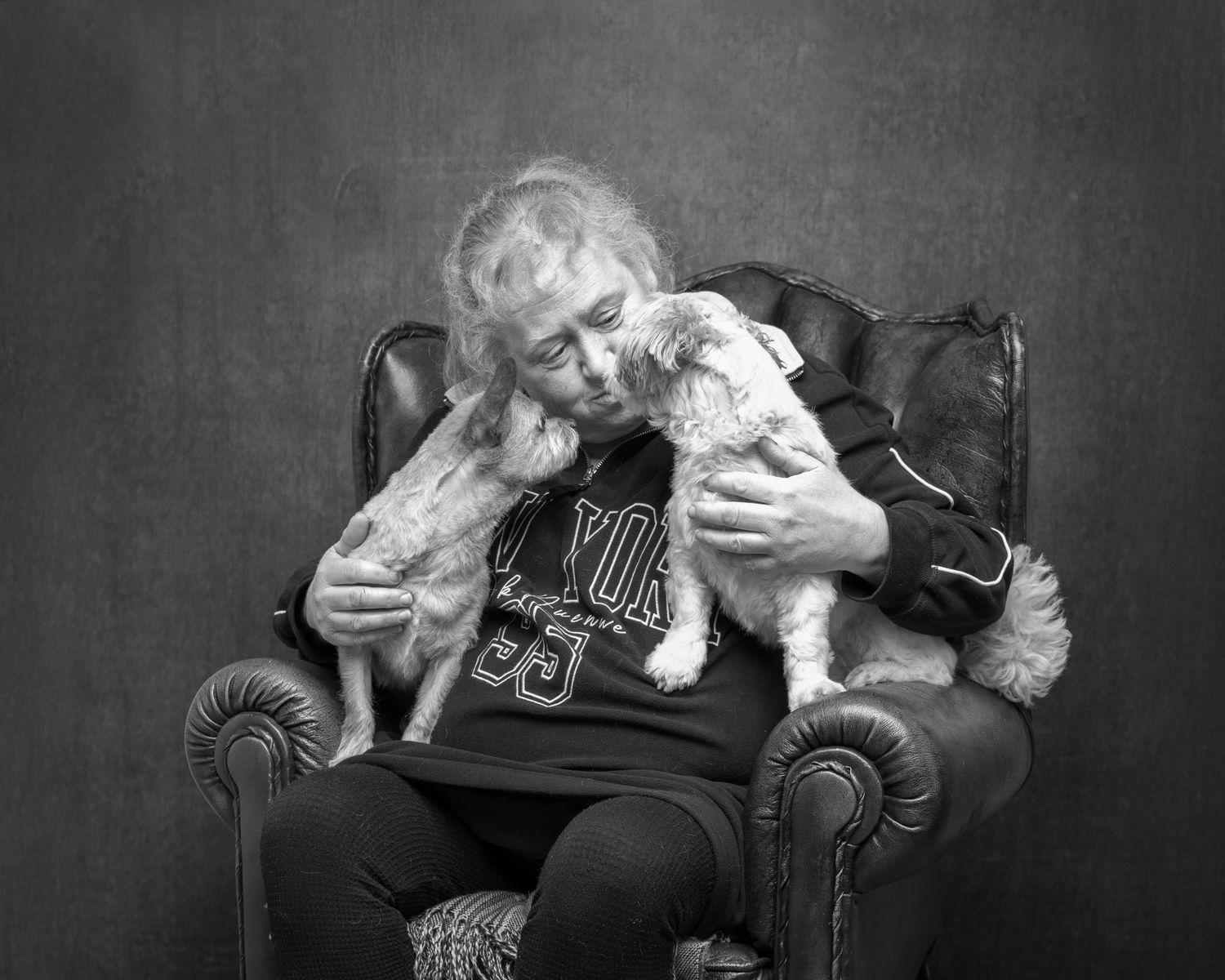 Elderly person sitting in leather armchair lovingly holding two small dogs in a black and white portrait photo.