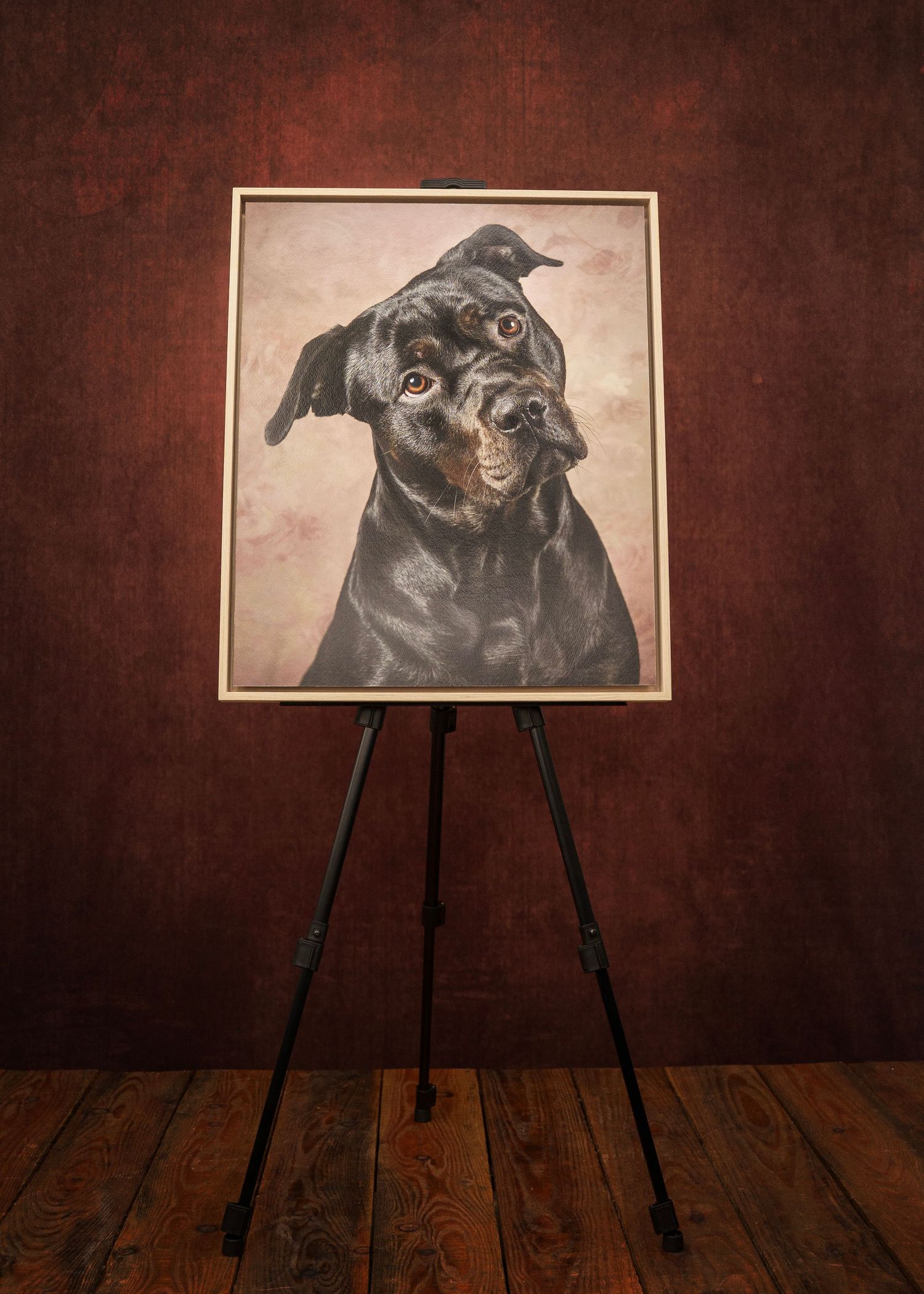 A framed portrait painting of a Rottweiler dog displayed on a black easel against a dark red wall.