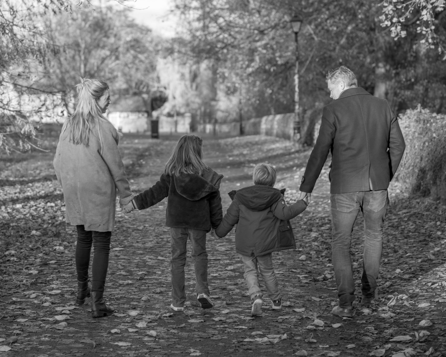 A family walks together holding hands along a leaf-covered path on an autumn day in black and white.