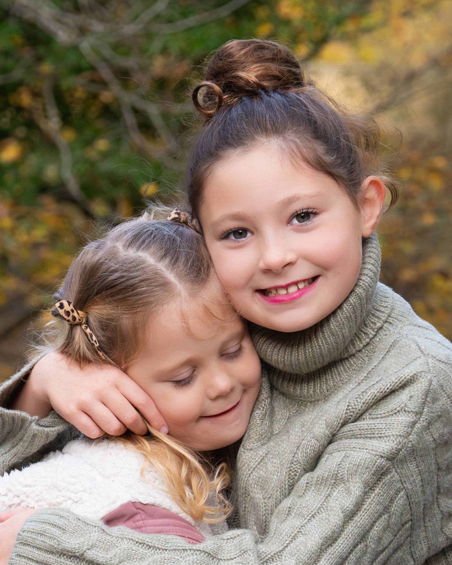 Two young sisters share a warm hug outdoors in autumn, wearing cozy green sweaters with colorful fall foliage behind them.