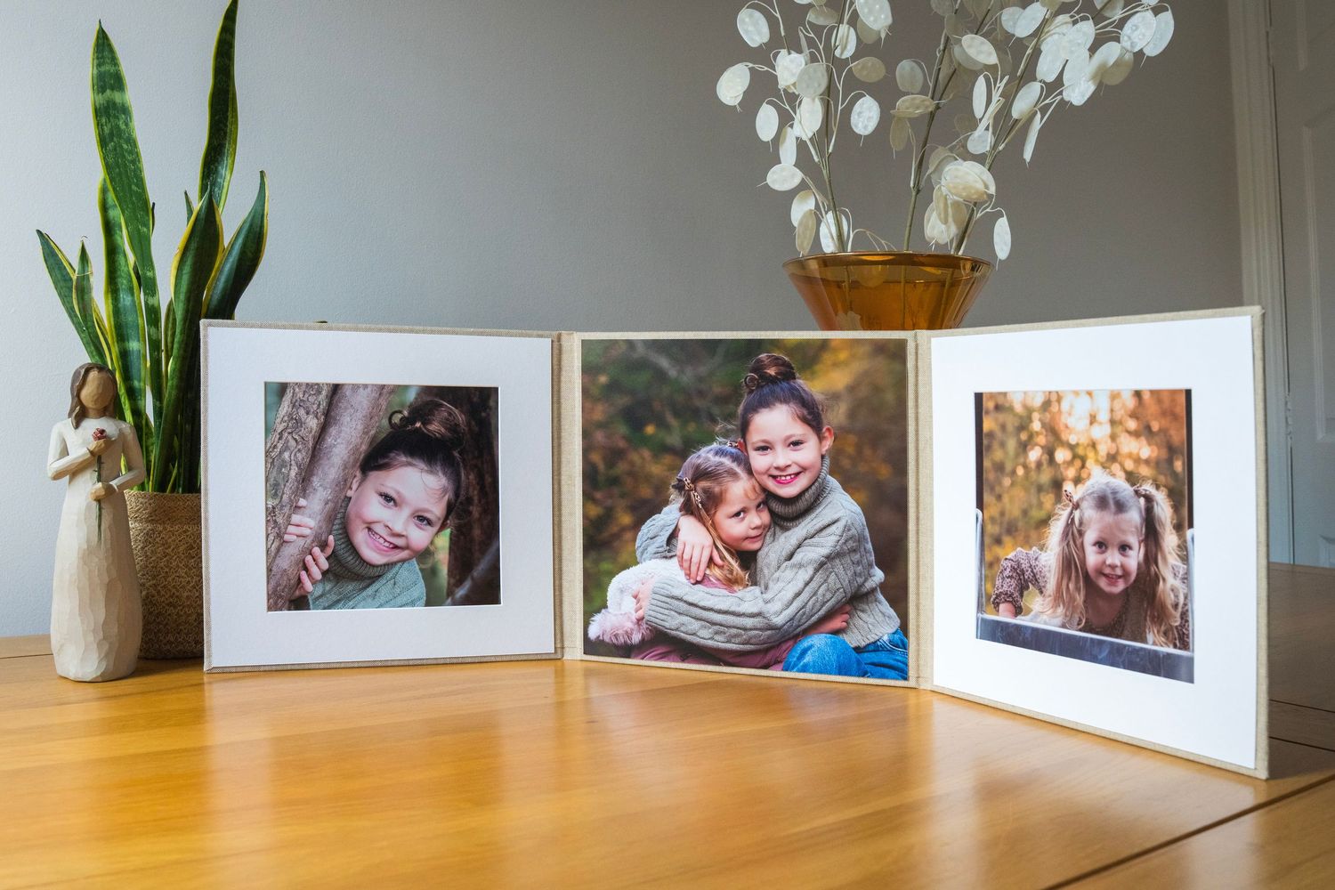 Three white photo frames displaying children portraits arranged on a wooden table with plants in the background.