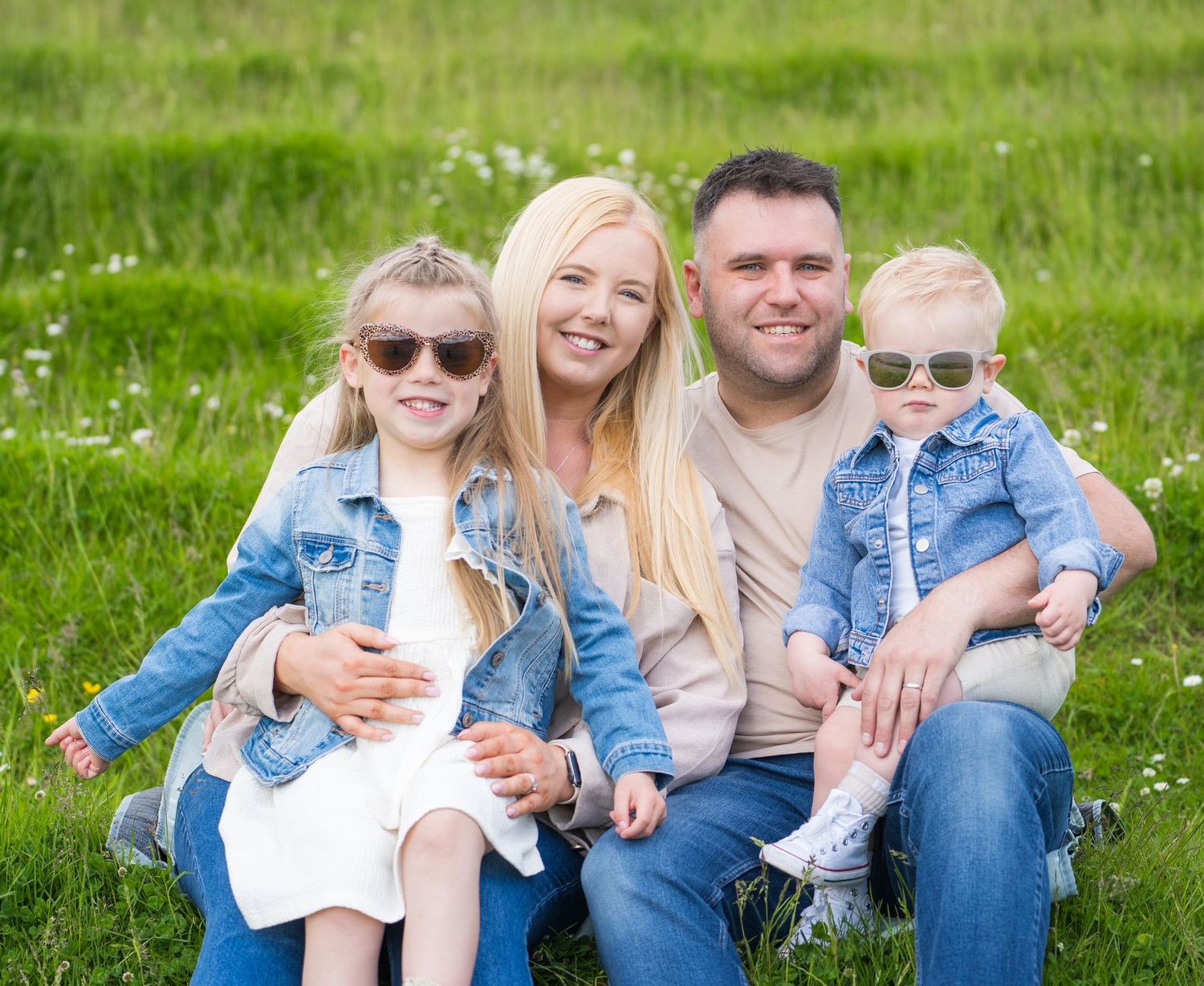 Happy family of four sitting in green field, parents and two children wearing denim jackets and sunglasses.