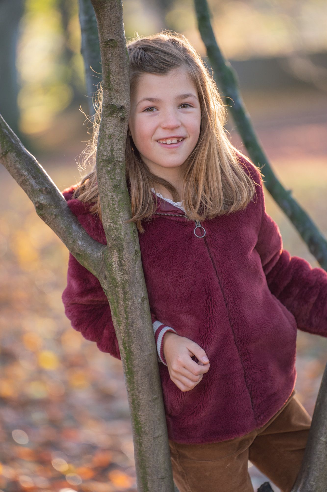 A child in a burgundy sweater poses playfully by a tree trunk in an autumn setting with golden bokeh background.