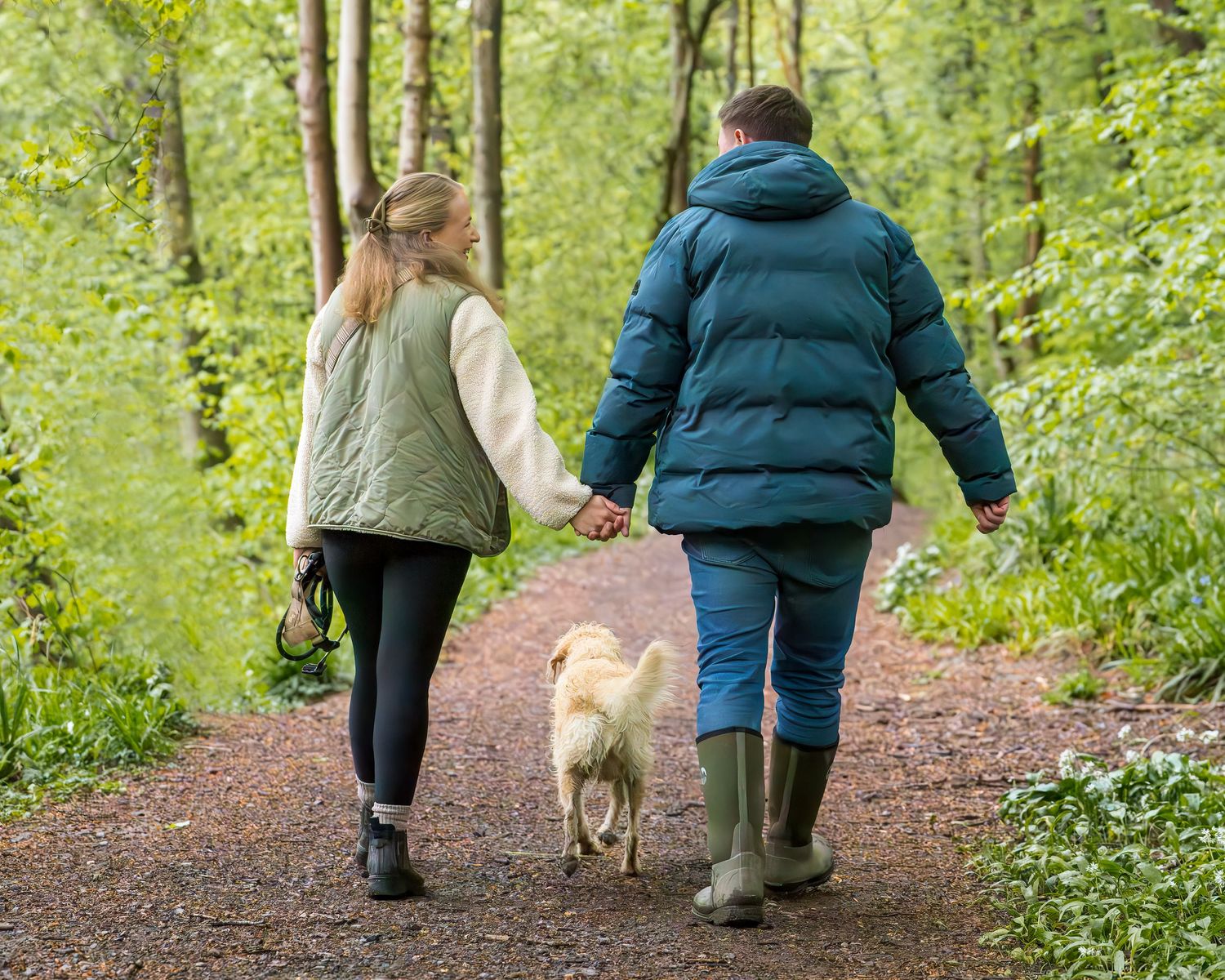 Two people holding hands walking a golden dog on a forest path, wearing puffer jackets and green wellies.