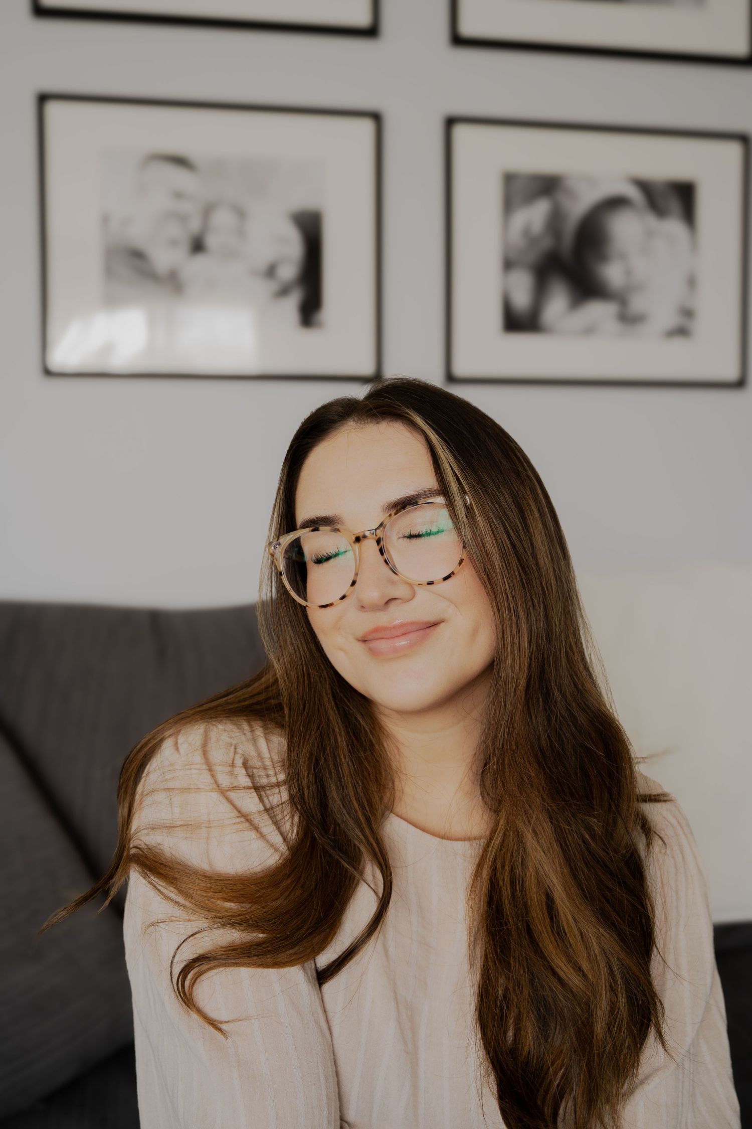 A person with long brown hair and glasses smiles while relaxing on a grey couch.